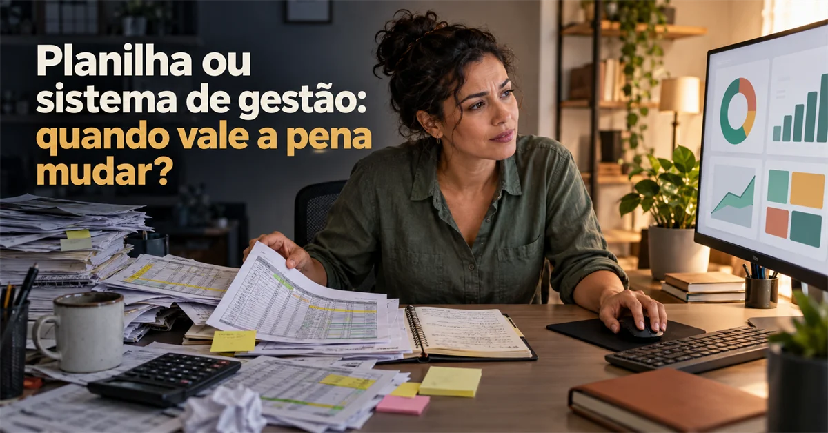 Mulher analisa pilhas de planilhas impressas na mesa enquanto o monitor ao lado exibe um painel de gestão com gráficos